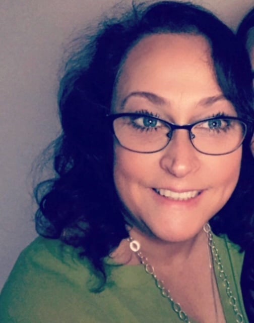 Woman with dark wavy hair and glasses wearing a green shirt, smiling at camera